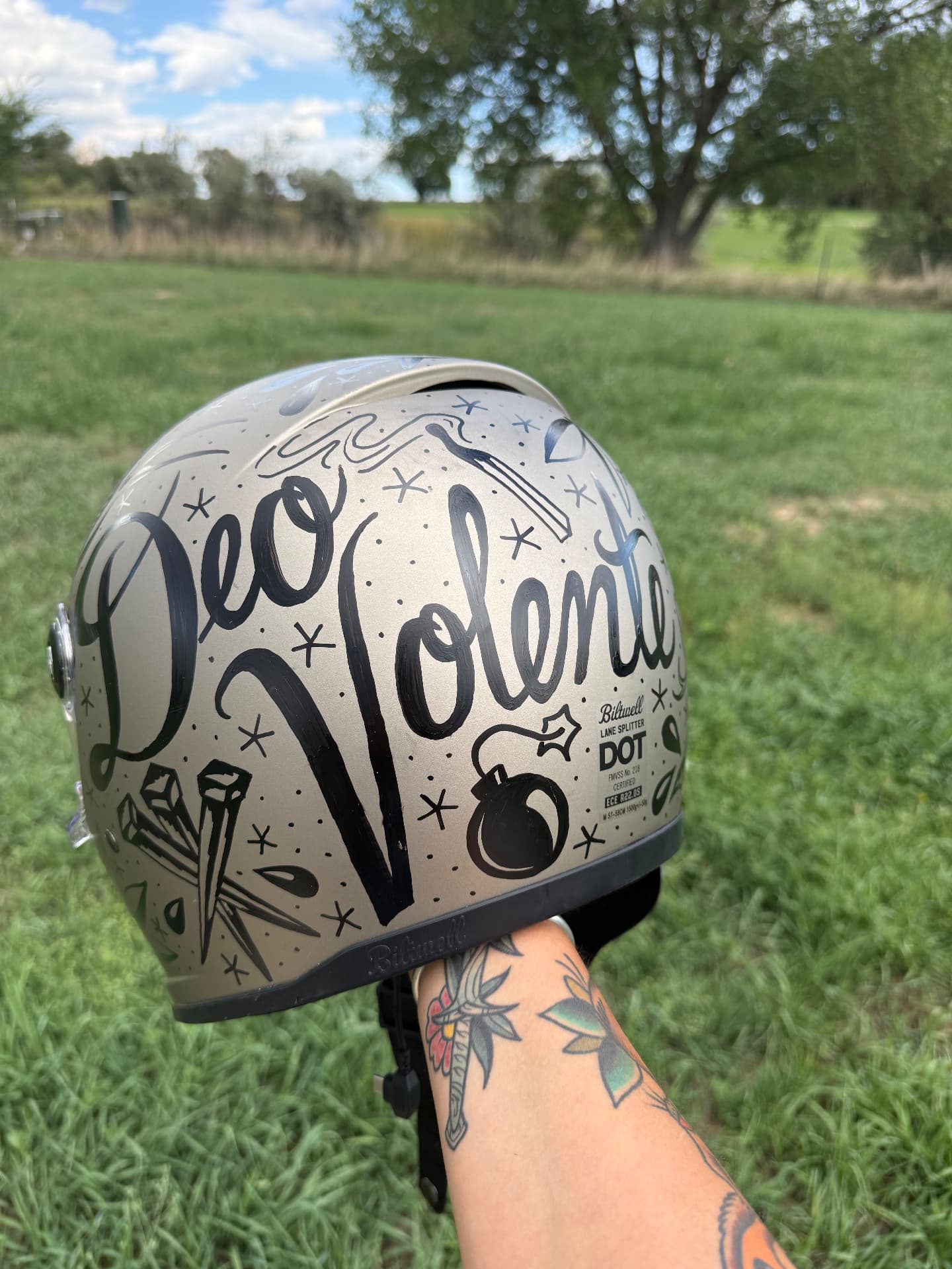 Rear of a hand-lettered helmet reading Deo Volente in black brush script with bombs, nails and stars